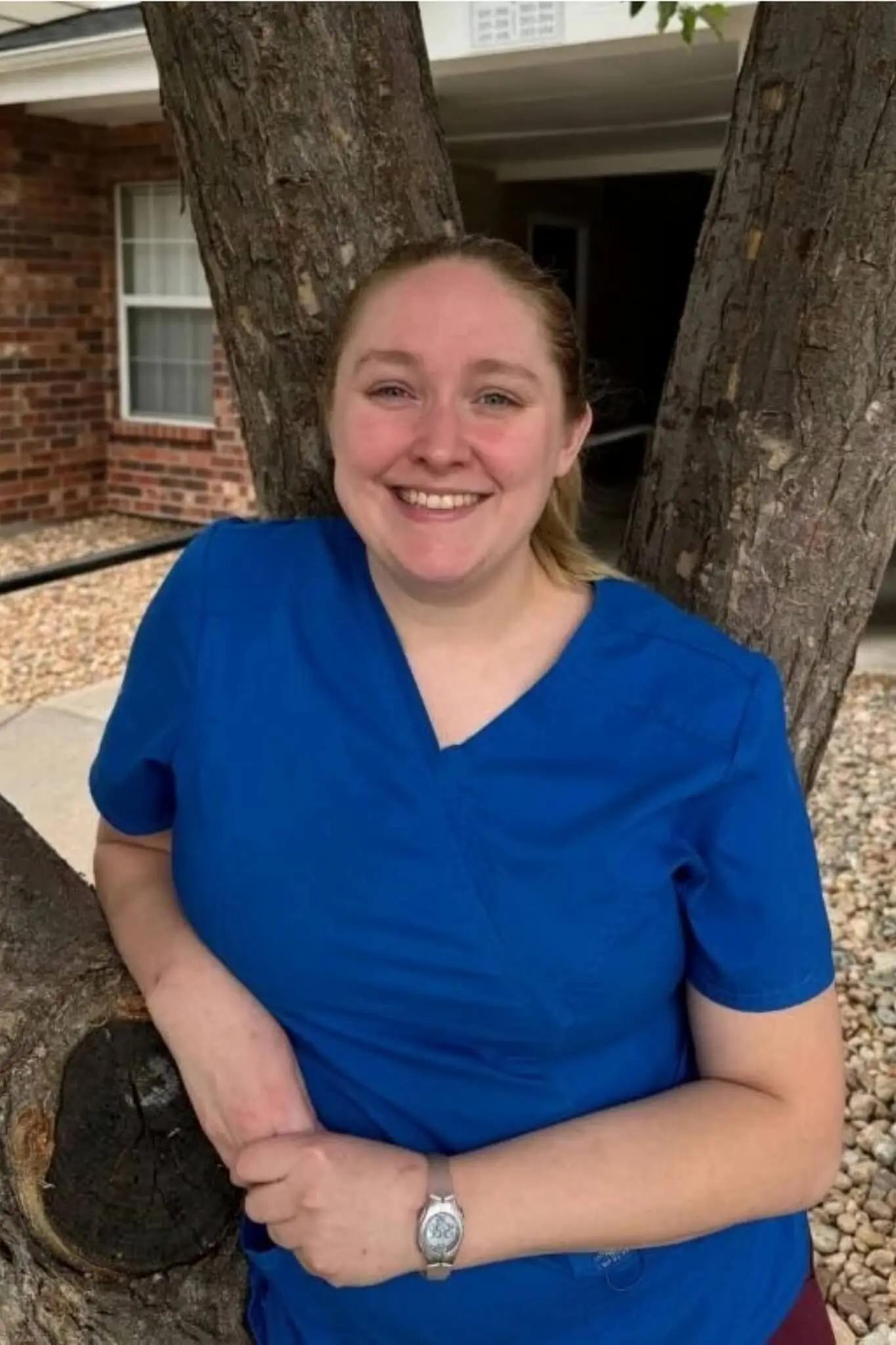 female wearing blue scrubs with blonde hair in a pony tail posing in front of a large tree
